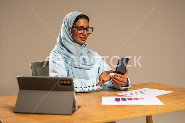 استخدام الهاتف المحمول في العمل أونلاين، مهن ووظائف مكتبية للنساء في السعودية، مفهوم العمل عن بعد عبر الانترنت، دمج الأعمال مع التكنولوجيا الحديثة، بورتريه لامرأة عربية خليجية سعودية محجبة تجلس خلف المكتب وأمامها الجهاز اللوحي وعدد من الأوراق، خلفية رمادية