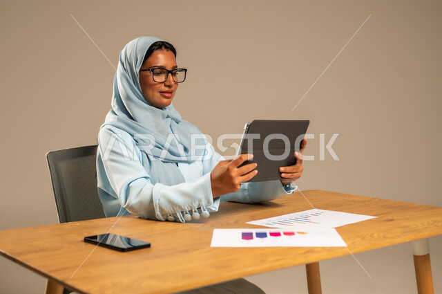 مفهوم العمل عن بعد عبر الانترنت، مهن ووظائف مكتبية إدارية نسائية في السعودية، إنجاز المهام المطلوبة أونلاين، دمج العمل مع التكنولوجيا الحديثة، بورتريه لامرأة عربية خليجية سعودية محجبة تجلس خلف المكتب وتقوم بمتابعة عملها عبر الجهاز اللوحي، خلفية رمادية