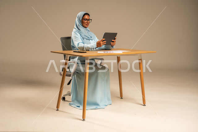 The concept of remote work via the Internet, female administrative office jobs and professions in Saudi Arabia, completing the required tasks online, integrating work with modern technology, a portrait of a veiled Saudi Arabian Gulf woman sitting behind a desk and following up on her work via a tablet, gray background