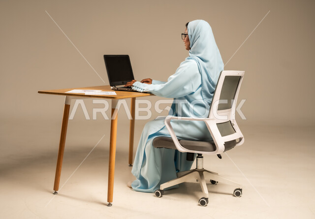 مهن مكتبية إدارية نسائية في السعودية، إنجاز المهام المطلوبة عبر جهاز الحاسوب المحمول، دمج العمل مع التكنولوجيا الحديثة، بورتريه من الخلف لامرأة عربية خليجية سعودية محجبة تجلس خلف المكتب وتقوم بمتابعة عملها أونلاين، مفهوم العمل عن بعد عبر الانترنت، خلفية رمادية