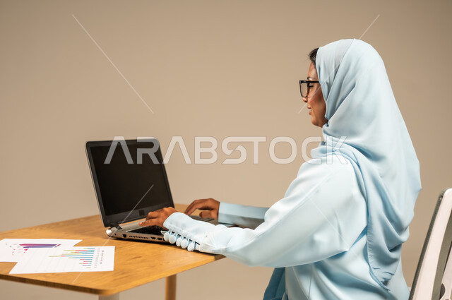 مهن مكتبية إدارية نسائية في السعودية، إنجاز المهام المطلوبة عبر جهاز الحاسوب المحمول، دمج العمل مع التكنولوجيا الحديثة، بورتريه من الخلف لامرأة عربية خليجية سعودية محجبة تجلس خلف المكتب وتقوم بمتابعة عملها أونلاين، مفهوم العمل عن بعد عبر الانترنت، خلفية رمادية