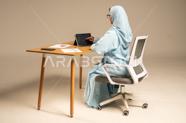 وظائف مكتبية إدارية نسائية في السعودية، إنجاز المهام المطلوبة أونلاين، دمج العمل مع التكنولوجيا الحديثة، بورتريه من الخلف لامرأة عربية خليجية سعودية محجبة تجلس خلف المكتب وتقوم بمتابعة عملها عبر الجهاز اللوحي، مفهوم العمل عن بعد عبر الانترنت، خلفية رمادية