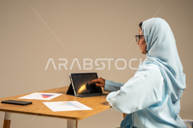 وظائف مكتبية إدارية نسائية في السعودية، إنجاز المهام المطلوبة أونلاين، دمج العمل مع التكنولوجيا الحديثة، بورتريه من الخلف لامرأة عربية خليجية سعودية محجبة تجلس خلف المكتب وتقوم بمتابعة عملها عبر الجهاز اللوحي، مفهوم العمل عن بعد عبر الانترنت، خلفية رمادية