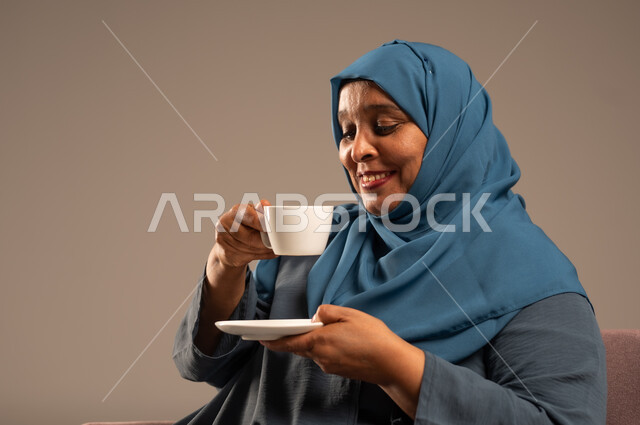  أصالة عادات وتقاليد المجتمع السعودي, كرم الضيافة وحسن الاستقبال في المملكة, بورتريه لامراة كبيرة بالسن عربية خليجية سعودية ترتدي العباءة والحجاب تجلس على كرسي مريح تحمل بيدها كوب من القهوة، الاهتمام بالمظهر الخارجي، خلفية ملونة