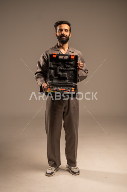 العمل في المجال الميكانيكي والكهربائي، أعمال الإصلاح وورشات الصيانة، بورتريه لعامل محترف عربي خليجي سعودي يرتدي اليونيفورم الخاص بالعمل يقف ويحمل حقيبة المعدات بين يديه بإيماءات البشاشة، مهن ووظائف للشباب السعودي، أدوات القياس والفحص اليدوية، خلفية رمادية
