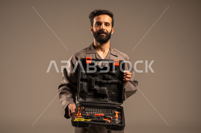 العمل في المجال الميكانيكي والكهربائي، أعمال الإصلاح وورشات الصيانة، بورتريه لعامل محترف عربي خليجي سعودي يرتدي اليونيفورم الخاص بالعمل يقف ويحمل حقيبة المعدات بين يديه بإيماءات البشاشة، مهن ووظائف للشباب السعودي، أدوات القياس والفحص اليدوية، خلفية رمادية