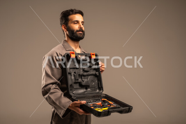 العمل في المجال الميكانيكي والكهربائي، أعمال الإصلاح وورشات الصيانة، بورتريه لعامل محترف عربي خليجي سعودي يرتدي اليونيفورم الخاص بالعمل يقف ويحمل حقيبة المعدات بين يديه بإيماءات البشاشة، مهن ووظائف للشباب السعودي، أدوات القياس والفحص اليدوية، خلفية رمادية