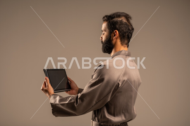 استخدام التقنيات الحديثة والتكنولوجيا في البحث والتطوير، أعمال الإصلاح والصيانة، بورتريه من الخلف لرجل عربي خليجي سعودي يرتدي اليونيفورم الخاص بالعمل يقف ويمسك الجاهز اللوحي بيديه، مهن ووظائف للشباب السعودي، العمل في المجال الميكانيكي والكهربائي، خلفية رمادية