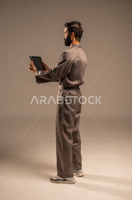 استخدام التقنيات الحديثة والتكنولوجيا في البحث والتطوير، أعمال الإصلاح والصيانة، بورتريه من الخلف لرجل عربي خليجي سعودي يرتدي اليونيفورم الخاص بالعمل يقف ويمسك الجاهز اللوحي بيديه، مهن ووظائف للشباب السعودي، العمل في المجال الميكانيكي والكهربائي، خلفية رمادية
