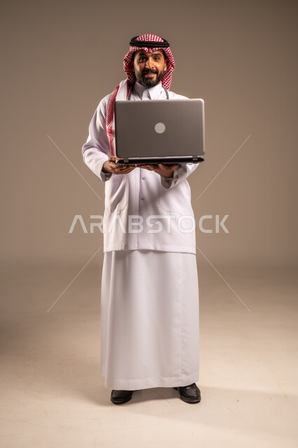استخدام الأجهزة التقنية الحديثة في الطب، بورتريه لطبيب عربي خليجي سعودي يرتدي الثوب التقليدي والشماغ والمعطف الأبيض يحمل بيده الحاسوب المحمول ينظر إلى الكاميرا، تقديم الاستشارات الطبية عن طريق الإنترنت، العمل في القطاع الصحي، الجسم بالطول الكامل، خلفية بيج