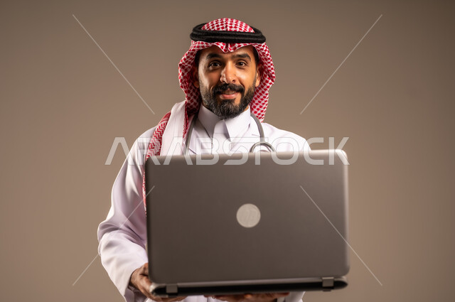 استخدام الأجهزة التقنية الحديثة في الطب، بورتريه مقرب لطبيب عربي خليجي سعودي يرتدي الثوب التقليدي والشماغ والمعطف الأبيض يحمل بيده الحاسوب المحمول ينظر إلى الكاميرا، تقديم الاستشارات الطبية عن طريق الإنترنت، العمل في قطاع الصحة والرعاية، خلفية بيج