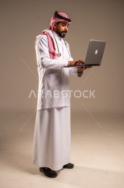 العمل في قطاع الصحة والرعاية، استخدام الأجهزة التقنية الحديثة في الطب، بورتريه من الجانب لطبيب عربي خليجي سعودي يرتدي الثوب التقليدي والشماغ والمعطف الأبيض يحمل بيده الحاسوب المحمول، تقديم الاستشارات الطبية عن طريق الإنترنت، الجسم بالطول الكامل، خلفية بيج