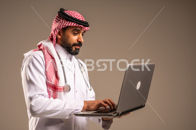 استخدام الأجهزة التقنية الحديثة في الطب، بورتريه مقرب من الجانب لطبيب عربي خليجي سعودي يرتدي الثوب التقليدي والشماغ والمعطف الأبيض يحمل بيده الحاسوب المحمول، تقديم الاستشارات الطبية عن طريق الإنترنت، العمل في قطاع الصحة والرعاية، خلفية بيج