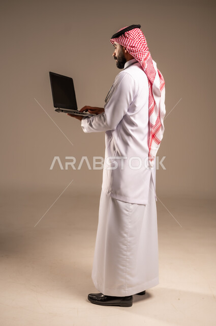 العمل في قطاع الصحة والرعاية، استخدام الأجهزة التقنية الحديثة في الطب، بورتريه من الجانب لطبيب عربي خليجي سعودي يرتدي الثوب التقليدي والشماغ والمعطف الأبيض يحمل بيده الحاسوب المحمول، تقديم الاستشارات الطبية عن طريق الإنترنت، الجسم بالطول الكامل، خلفية بيج