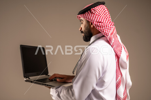 Working in the health sector in Saudi Arabia, using modern technical devices in medicine, close-up side portrait of a Saudi Arabian Gulf doctor wearing traditional thobe, shemagh and white coat holding a laptop, providing medical consultations via the internet, beige background
