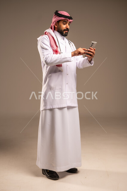 العمل في قطاع الصحة والرعاية، البرامج والتطبيقات الطبية، استخدام الأجهزة التقنية الحديثة، بورتريه لطبيب عربي خليجي سعودي يرتدي الثوب التقليدي والشماغ والمعطف الأبيض يضع السماعة الطبية حول عنقه يستخدم الهاتف المحمول، الجسم بالطول الكامل، خلفية بيج