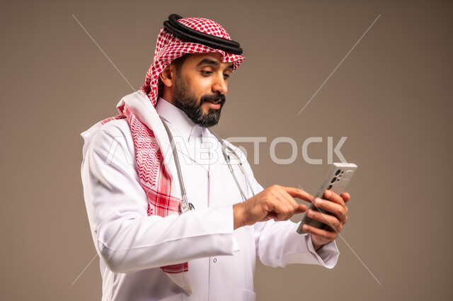العمل في قطاع الصحة والرعاية في المملكة العربية السعودية، البرامج والتطبيقات الطبية، استخدام الأجهزة التقنية الحديثة، بورتريه مقرب لطبيب عربي خليجي سعودي يرتدي الثوب التقليدي والشماغ والمعطف الأبيض يضع السماعة الطبية حول عنقه يستخدم الهاتف المحمول، خلفية بيج