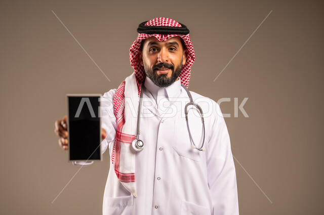 البرامج والتطبيقات الطبية، استخدام الأجهزة التقنية الحديثة، عرض شاشة سوداء فارغة، بورتريه مقرب لطبيب عربي خليجي سعودي يرتدي الثوب التقليدي والشماغ والمعطف الأبيض يضع السماعة الطبية حول عنقه يحمل بيده الهاتف المحمول، تقديم الاستشارات عن طريق الإنترنت، خلفية بيج
