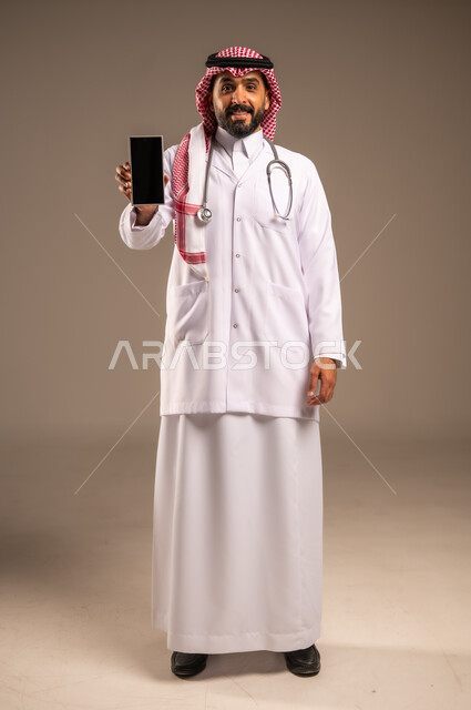 Medical programs and applications, use of modern technical devices, display of a blank black screen, portrait of a Saudi Arabian Gulf doctor wearing a traditional thobe, shemagh and white coat, wearing a stethoscope around his neck, holding a mobile phone in his hand, full body, beige background