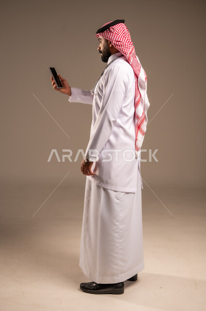 Working in the health sector, using modern technical devices in medicine, side portrait of a Saudi Arabian Gulf doctor wearing traditional thobe, shemagh and white coat holding a mobile phone, providing medical consultations via the internet, full body, beige background