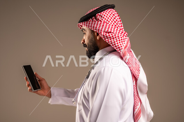 العمل في القطاع الصحي، استخدام الأجهزة التقنية الحديثة في الطب، بورتريه مقرب من الجانب لطبيب عربي خليجي سعودي يرتدي الثوب التقليدي والشماغ والمعطف الأبيض يحمل بيده الهاتف المحمول، تقديم الاستشارات الطبية عن طريق الإنترنت، خلفية بيج