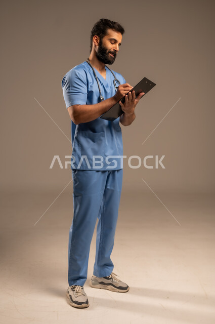 إعداد التقارير وتقديم الخدمات الصحية، بورتريه لممرض عربي خليجي سعودي يرتدي اليونيفورم يضع السماعة الطبية حول عنقه يحمل بيده ملفات المرضى، العمل في القطاع الصحي في المملكة العربية السعودية، حسن التعامل مع المراجعين، الجسم بالطول الكامل، خلفية بيج