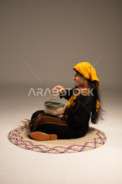Anniversary of the founding of the first Saudi state February 22, Saudi national local products and crops, the day we started 1727 AD, close-up portrait from the side of a smiling Saudi Arabian Gulf girl wearing a shayla and a sidra putting grains in a basket, sitting on a straw mat, wearing the costumes of the southern region, beige background