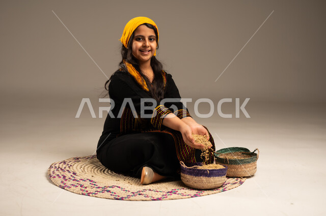 الجلوس على حصيرة مصنوعة من القش، منتجات ومحاصيل محلية وطنية سعودية، ذكرى تأسيس الدولة السعودية الأولى 22 فبراير، يوم بدينا 1727 م، بورتريه مقرب لفتاة عربية خليجية سعودية مبتسمة ترتدي الشيلة والسدره تقوم بوضع الحبوب في السلة، ، ارتداء أزياء المنطقة الجنوبية، مهن وحرف يدوية شعبية قديمة، خلفية بيج