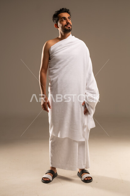 زيارة البيت الحرام في مكة المكرمة، مفهوم العبادة والتقرب الى الله، بورتريه مقرب لرجل عربي خليجي سعودي مبتسم يرتدي ثوب الاحرام ينظر الى مكان ما بإيماءات السعادة والطمأنينة، أداء مناسك الحج والعمرة، الركن الخامس من أركان الاسلام لمن استطاع اليه سبيلا، الجسم الكامل بالطول، خلفيه بيج