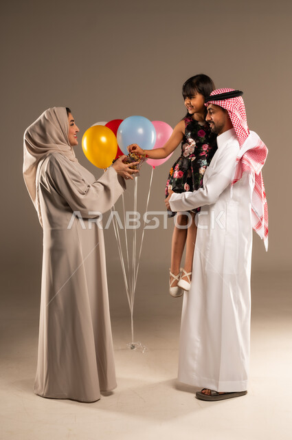 الألفة والمودة بين أفراد الأسرة السعودية, بورتريه لرجل عربي خليجي سعودي يحمل ابنته بايماءات السعادة, امرأة عربية خليجية محجبة تقوم بتقديم الحلويات للأقارب في العيد المبارك,  ايماءات الرضا والسرور، خلفية بيج