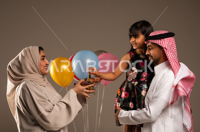 الألفة والمودة بين أفراد الأسرة السعودية, بورتريه لرجل عربي خليجي سعودي يحمل ابنته بايماءات السعادة, امرأة عربية خليجية محجبة تقوم بتقديم الحلويات للأقارب في العيد المبارك,  ايماءات الرضا والسرور، خلفية بيج