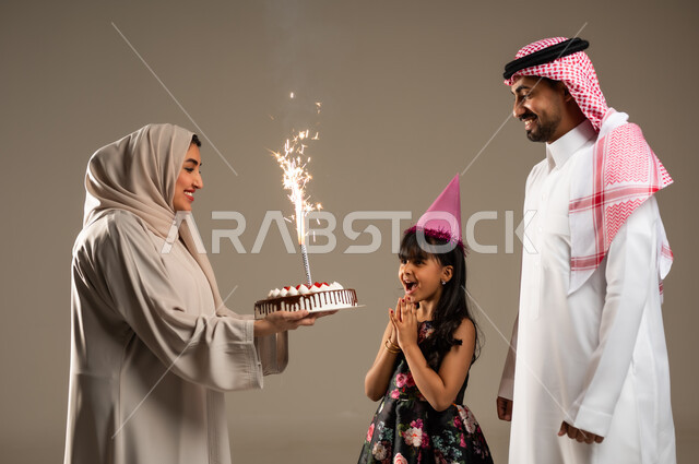 ايماءات البهجة والسرور, الاجتماعات والاحتفالات العائلية، بورتريه مقرب لعائلة عربية خليجية سعودية يحتفلون بعيد ميلاد طفلتهم ويقدمون لها قالب الكيك المزين، الألفة والمودة بين أفراد الأسرة السعودية، خلفية بيج