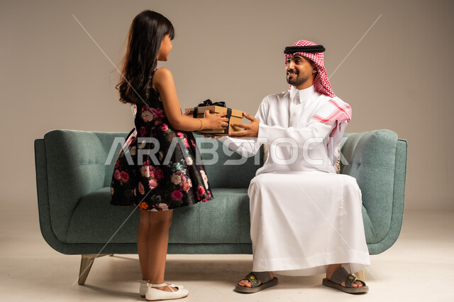 الألفة والمودة بين أفراد الأسرة السعودية, بورتريه من الخلف لطفلة عربية خليجية سعودية تقدم الهدية لوالدها بايماءات الرضا والسرور, تقديم الهدايا بالمناسبات وأعياد الميلاد, خلفية بيج