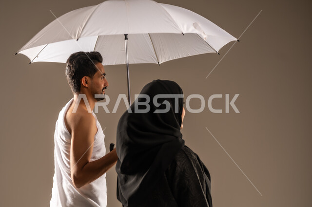  زيارة بيت الله الحرام، استخدام المظلة الشمسية للحماية والتظلل،  الاستعداد لأداء مناسك الحج والعمرة، بورتريه مقرب من الخلف لزوجين عربيين خليجيين سعوديين يرتديان ثياب الاحرام ينظران الى مكان ما، مفهوم العبادة والتقرب الى الله تعالى، خلفية بيج