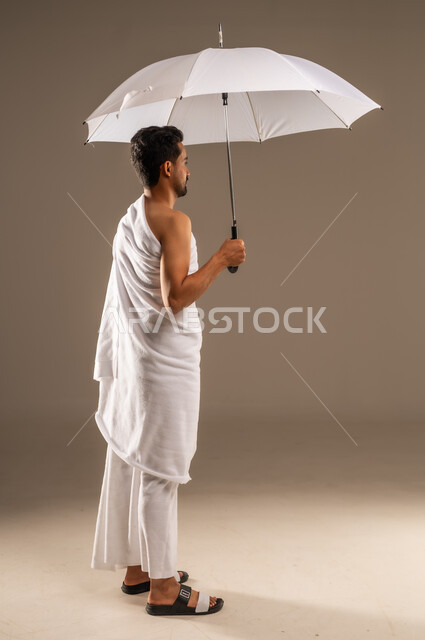 العبادة والتقرب الى الله بالأعمال الصالحة، أداء مناسك فريضة الحج و العمرة، زيارة بيت الله الحرام في مكة المكرمة، بورتريه مقرب من الخلف لرجل مسلم عربي خليجي سعودي يرتدي ثياب الاحرام يحمل بيده مظلة شمسية بيضاء للحماية والتظلل، الجسم الكامل بالطول، خلفية بيج