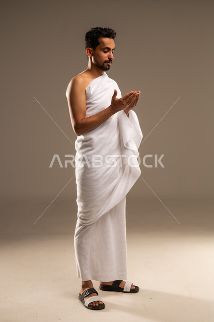 Maintaining prayers, supplications and asking forgiveness from Allah Almighty, visiting the Holy House in Mecca, the concept of worship and drawing closer to Allah, a close-up portrait of a Saudi Arabian Gulf Arab Muslim man wearing the Ihram garment, supplicating with humility and supplication, performing the rituals of Hajj and Umrah, full body length, beige background