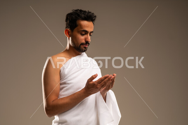 Maintaining prayers, supplications and asking forgiveness from Allah Almighty, visiting the Holy House in Mecca, the concept of worship and drawing closer to Allah, a close-up portrait of a Saudi Arabian Gulf Arab Muslim man wearing the Ihram garment, supplicating with humility and supplication, performing the rituals of Hajj and Umrah, beige background