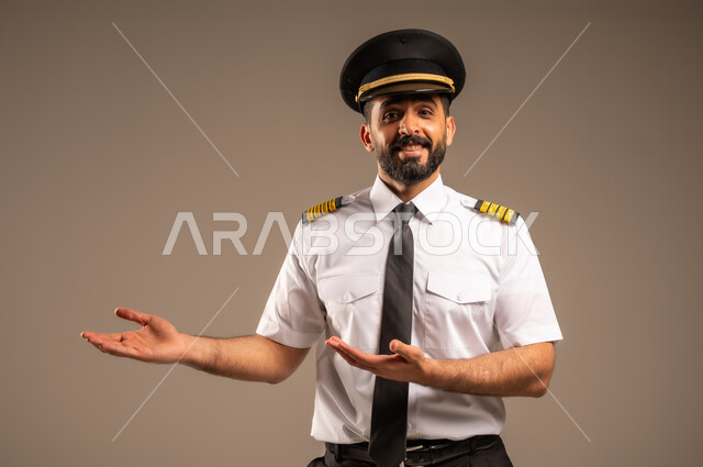 خطوط الطيران السعودية, طاقم الاستقبال والترحيب بالركاب، العمل في مجال الطيران، مفهوم السياحة والسفر، رحلات جوية سياحية, بورتريه مقرب لكابتن طيار عربي سعودي خليجي مبتسم يرتدي الزي الرسمي يشير بيديه الى الجانب بتعابير السعادة والسرور، خلفية رمادية