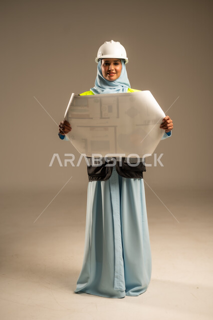 الإشراف على المشاريع الهندسية في موقع العمل, تطوير و بناء المملكة العربية السعودية على يد ابنائها, مهن ووظائف هندسية نسائية, بورتريه مقرب لمهندسة عربية خليجية سعودية محجبة ترتدي خوذة الحماية و السترة تحمل بيدها مخطط البناء، خلفية رمادية
