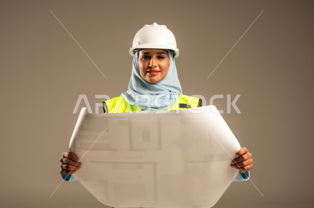 Supervising engineering projects on the work site, developing and building the Kingdom of Saudi Arabia by its sons, female engineering professions and jobs, close-up portrait of a veiled Saudi Arabian Gulf female engineer wearing a protective helmet and jacket holding a construction plan in her hand, gray background