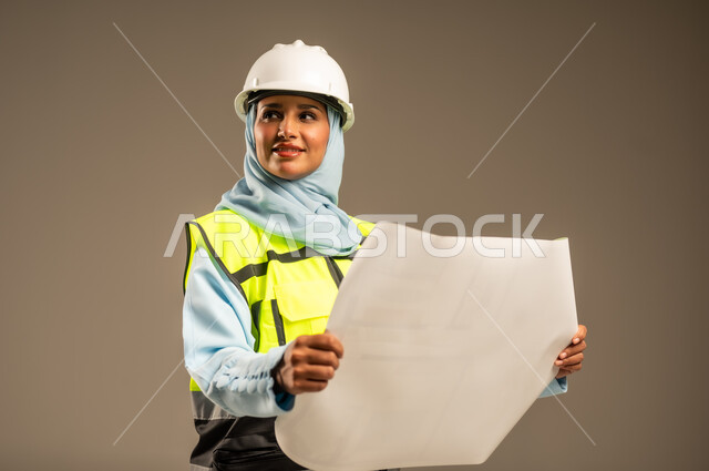 Supervising engineering projects on the work site, developing and building the Kingdom of Saudi Arabia by its sons, female engineering professions and jobs, close-up portrait of a veiled Saudi Arabian Gulf female engineer wearing a protective helmet and jacket holding a construction plan in her hand, gray background