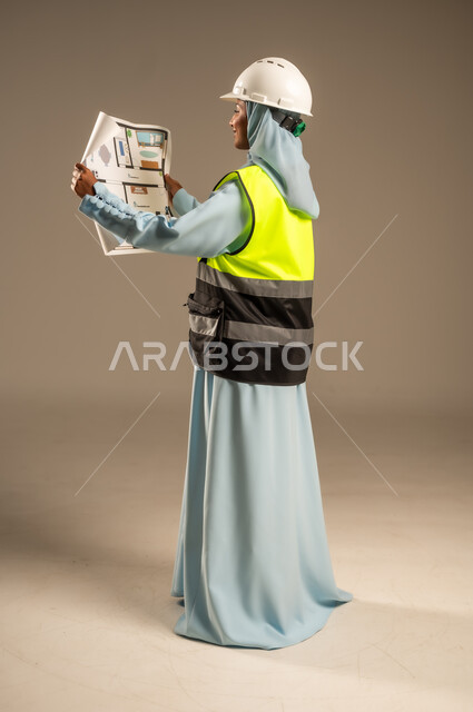 تطوير و بناء المملكة العربية السعودية على يد ابنائها, مهن ووظائف هندسية نسائية, بورتريه لمهندسة عربية خليجية سعودية محجبة ترتدي خوذة الحماية و السترة تحمل بيدها مخطط البناء، الإشراف على المشاريع الهندسية في موقع العمل, الجسم بالطول الكامل, خلفية رمادية