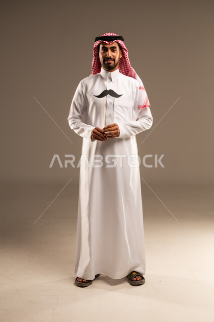 اليوم العالمي للرجل 19 نوفمبر، مفهوم القوة والرجولة، النظر الى الكاميرا بإيماءات الفخر والإعتزاز بالنفس، بورتريه لشاب عربي خليجي سعودي يرتدي الثوب التقليدي والشماغ يمسك بيده عود خشبي مع قصاصة ورقية على شكل شارب، صورة الجسم بالطول الكامل, خلفية بني