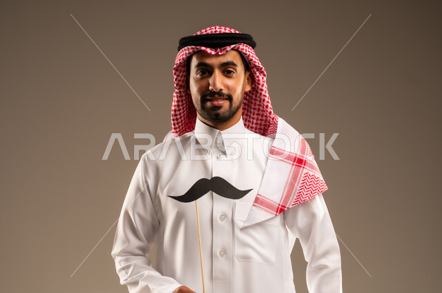 اليوم العالمي للرجل 19 نوفمبر، مفهوم القوة والرجولة، إيماءات الفخر والإعتزاز بالنفس، الإهتمام بصحة الذكور، بورتريه لشاب عربي خليجي سعودي يرتدي الثوب التقليدي والشماغ يمسك بيده عود خشبي مع قصاصة ورقية على شكل شارب، خلفية بني