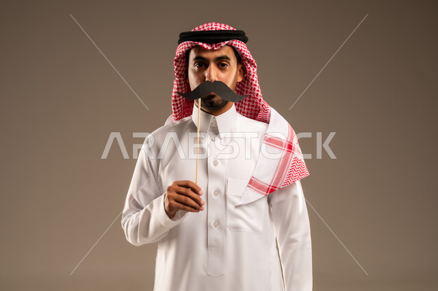 ذكرى اليوم العالمي للرجل 19 نوفمبر، الوقوف باستقامة والنظر إلى الكاميرا بإيماءات الحزم والجدية، بورتريه مقرب لشاب عربي خليجي سعودي يرتدي الزي التقليدي و الشماغ يمسك بيده عود خشبي مع قصاصة ورقية على شكل شارب ويضعها على وجهه، خلفية بني