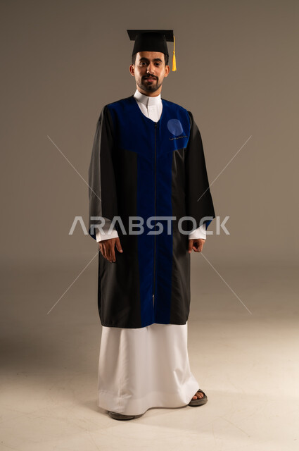  الشعور بالفرح والفخر والثقة بالنفس, مفهوم الاجتهاد والتفوق، الاحتفال بإنهاء الدراسة الجامعية، بورتريه لطالب عربي سعودي خليجي يرتدي العباءه وقبعة التخرج ينظر إلى الكاميرا بإيماءات الرضا والسرور, صورة الجسم بالطول الكامل, خلفية بنية 