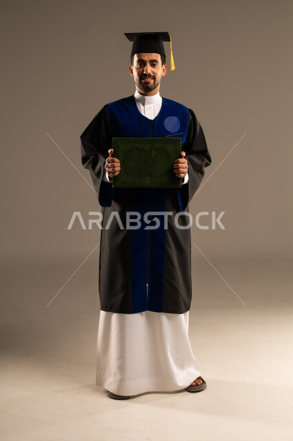  الشعور بالفرح والفخر والثقة بالنفس, مفهوم الاجتهاد والتفوق، الاحتفال بإنهاء الدراسة الجامعية، بورتريه لطالب عربي سعودي خليجي يرتدي العباءه وقبعة التخرج ينظر إلى الكاميرا يحمل بيده الاطروحة بإيماءات الرضا والسرور, صورة الجسم بالطول الكامل, خلفية بنية 