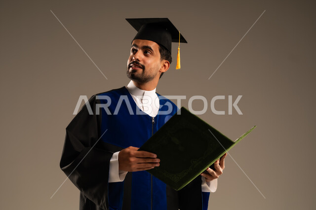 الاحتفال بإنهاء الدراسة الجامعية، بورتريه مقرب لطالب عربي سعودي خليجي يرتدي العباءه وقبعة التخرج ينظر إلى شيء ما يحمل بيده الاطروحة بإيماءات الرضا والسرور, الشعور بالفرح والفخر والثقة بالنفس, مفهوم الاجتهاد والتفوق، خلفية بنية