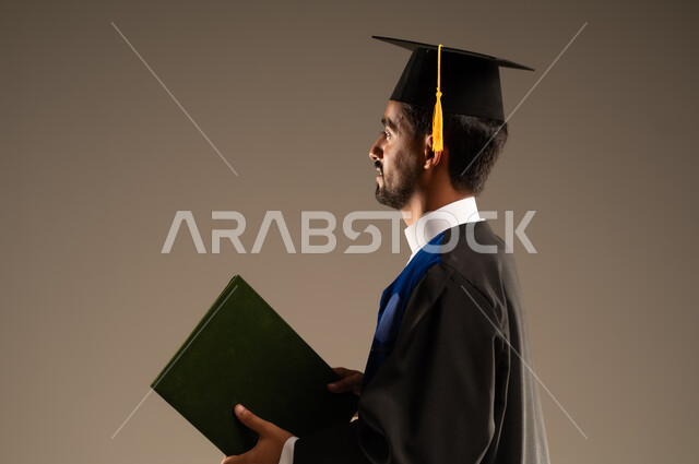 ايماءات تدل على الشعور بالفخر والسعاده, بورتريه مقرب من الجانب لطالب عربي سعودي خليجي يرتدي العباءه وقبعة التخرج يمسك بيده شهادة التفوق والنجاح, الاحتفال بإنهاء الدراسة الجامعية, خلفية بنية