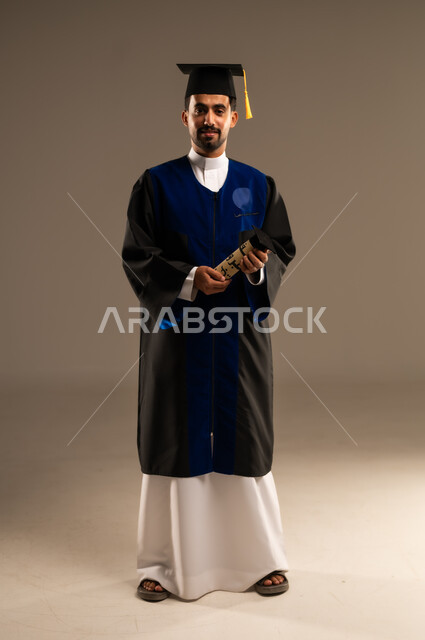 النظر الى الكاميرا بايماءات تدل على الشعور بالفخر والسعاده, بورتريه لطالب عربي سعودي خليجي يرتدي العباءه وقبعة التخرج يمسك بيده شهادة التفوق والنجاح, الاحتفال بإنهاء الدراسة الجامعية، صورة الجسم بالطول الكامل, خلفية بنية